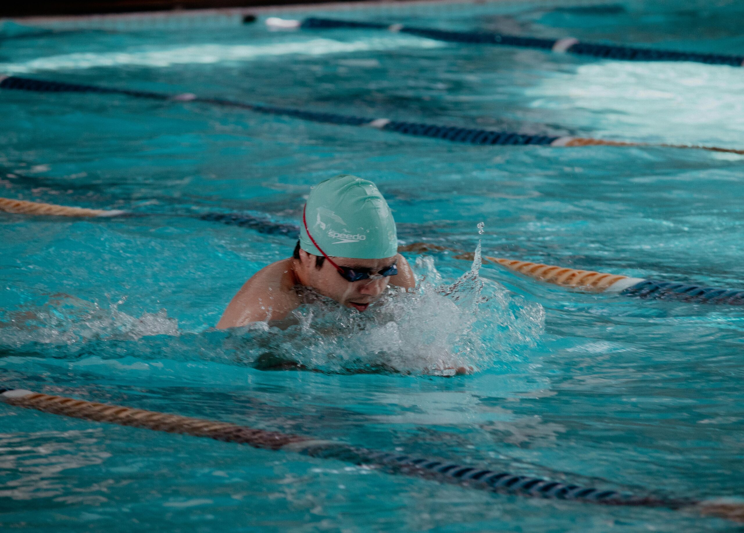 Swimmer doing butterfly stroke in indoor pool, wearing goggles and cap. Intense training session.