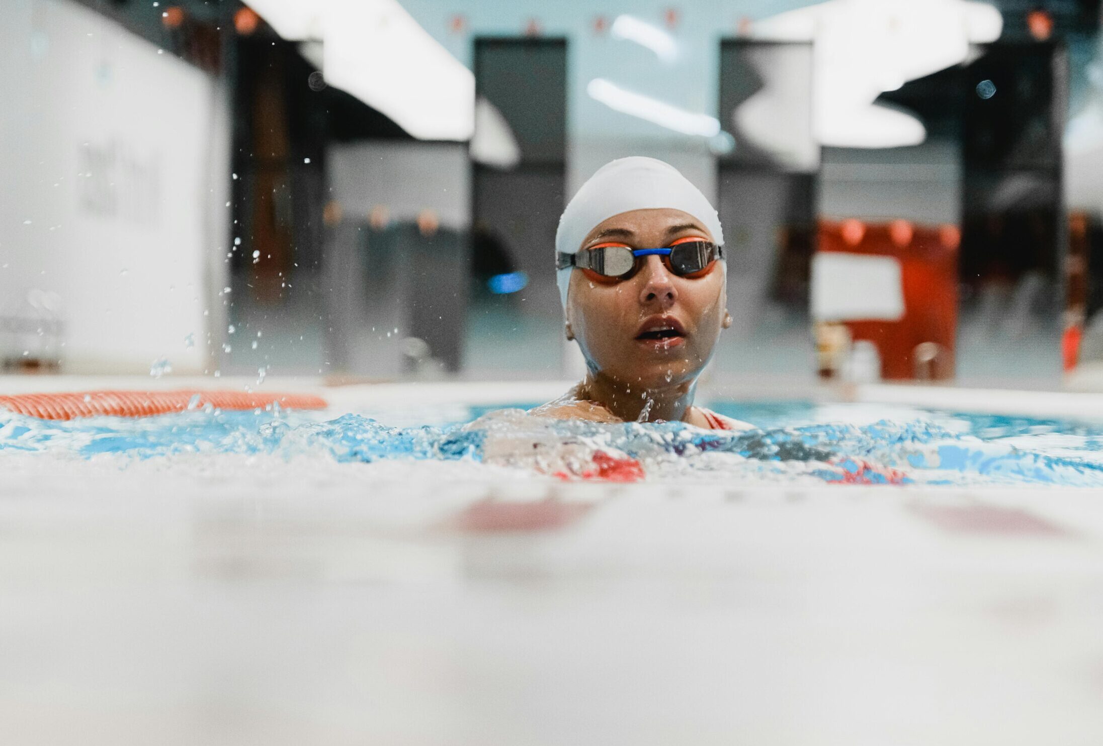 A focused female swimmer in an indoor pool, wearing goggles and swim cap, captured in mid-stroke.